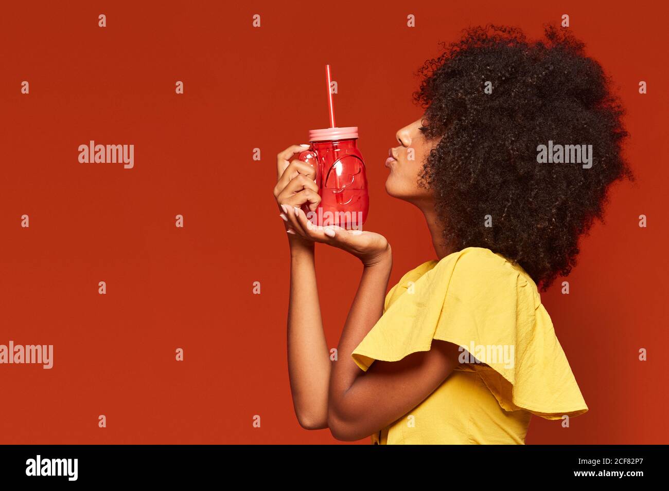 Cheerful young African American lady with curly hair holding red jar with straw and enjoying beverage on red background doing a kiss gesture Stock Photo