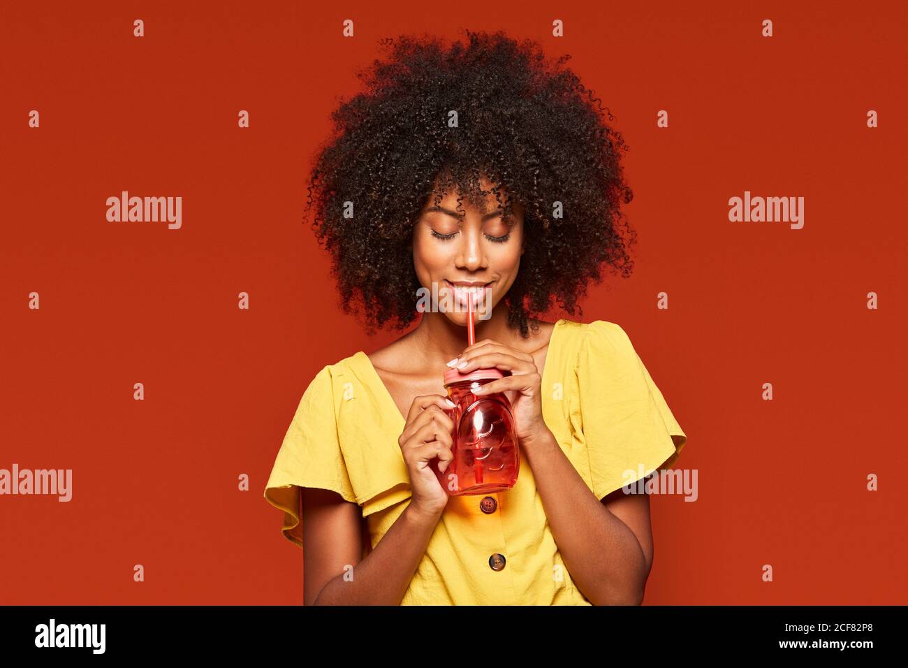 Dreamy young African American lady with curly hair holding red jar with straw and enjoying beverage on red background Stock Photo