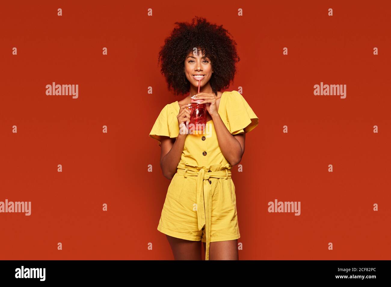 Cheerful young African American lady with curly hair holding red jar with straw and enjoying beverage on red background looking at camera Stock Photo