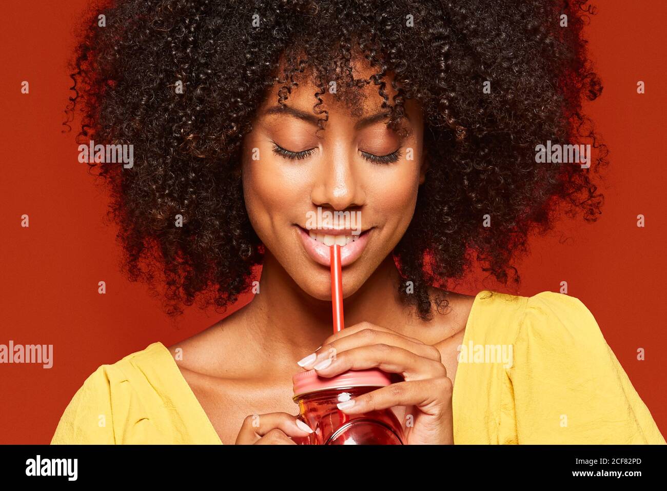Close-up view of dreamy young African American lady with curly hair holding red jar with straw and enjoying beverage on red background Stock Photo