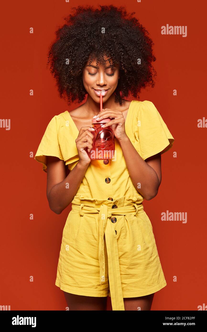 Dreamy young African American lady with curly hair holding red jar with straw and enjoying beverage on red background Stock Photo