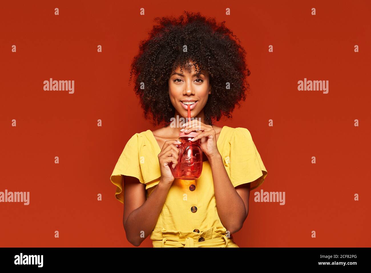 Dreamy young African American lady with curly hair holding red jar with straw and enjoying beverage on red background looking at camera Stock Photo