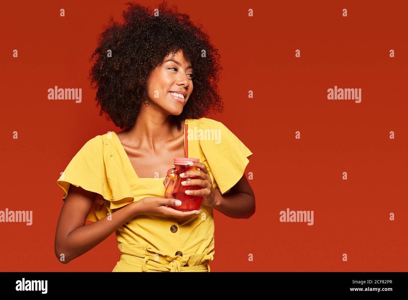 Cheerful young African American lady with curly hair holding red jar with straw and enjoying beverage on red background Stock Photo