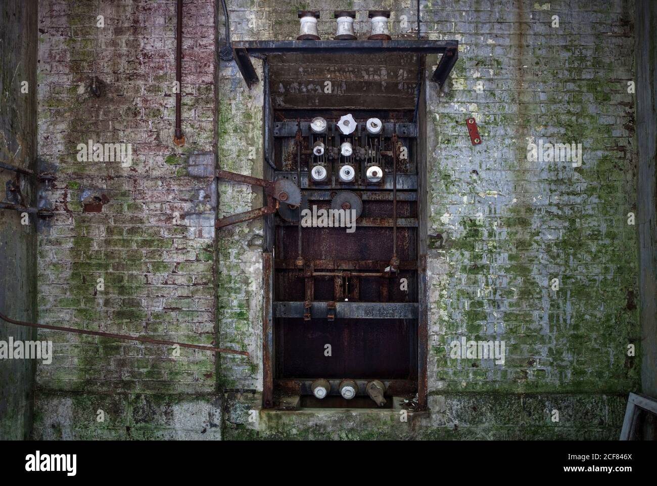 Old broken rusty mechanism with worn steel pipes of coils and rods in dilapidated gray brick wall covered with moss in abandoned factory Stock Photo