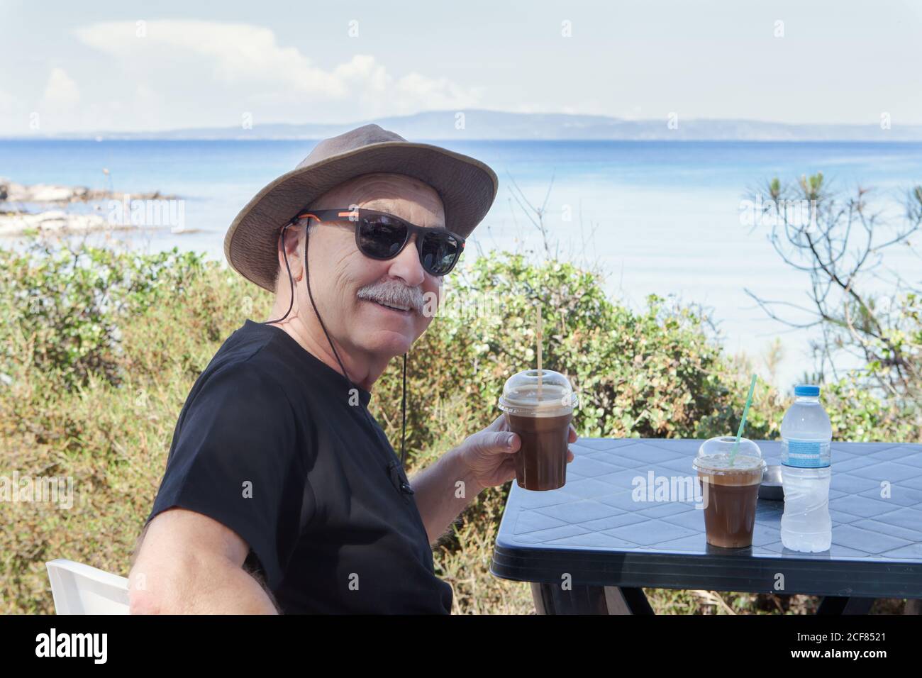 Smiling old man in sunglasses and hat holding plastic glass of drink and looking at camera sitting at table on seaside in Halkidiki, Greece Stock Photo