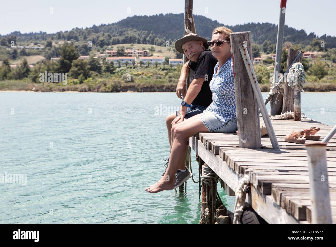 Relaxing elderly man and Woman in hats and sunglasses enjoying time together while sitting on pier and looking at camera in Halkidiki, Greece Stock Photo