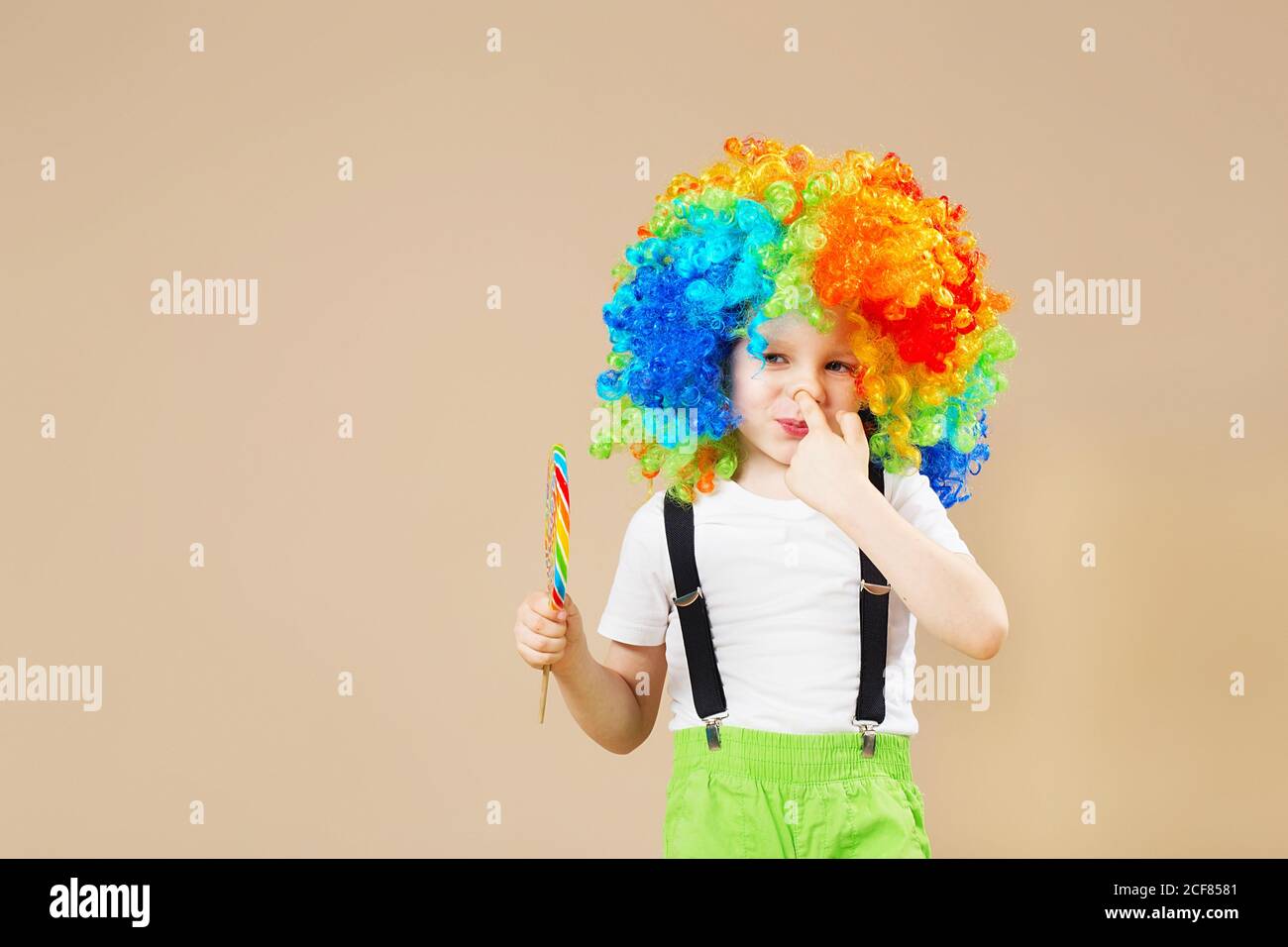 Happy clown boy in large colorful wig. Let's party! Funny kid clown. 1 April Fool's day concept. child eating lollipop. Finger in the nose. Picking in Stock Photo