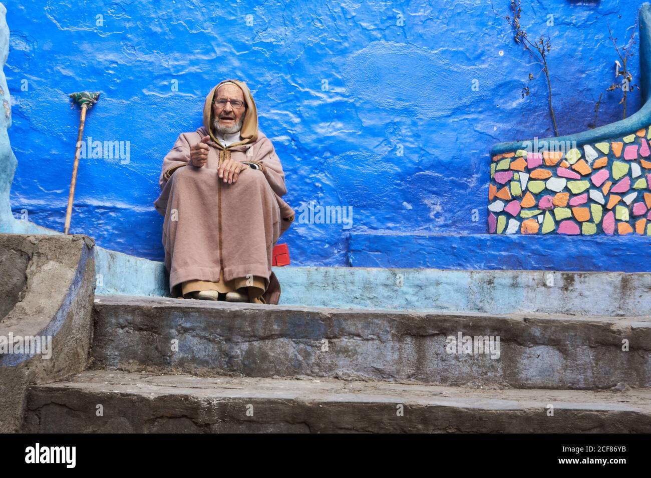 Marrakesh, Morocco - 31 December 2017: Senior adult man in cape sitting on step at blue wall and looking at camera Stock Photo