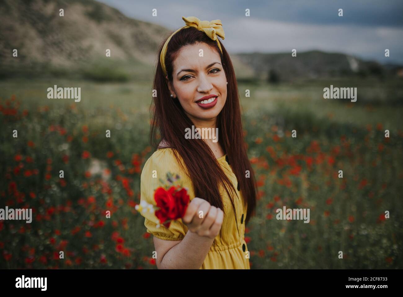 Smiling attractive red haired adult female in yellow dress and headband with red lips holding bouquet with red rose and looking at camera while standing alone in blurred green meadow with red flowers against hills under gray cloudy sky during daytime Stock Photo
