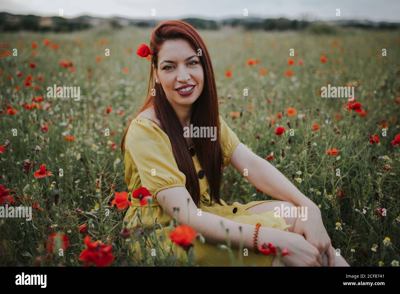Pensive attractive red haired adult lady in yellow dress with red poppy in hair and red lips looking over shoulder at camera while sitting alone in blurred amazing green meadow with red and white flowers against hills under gray cloudy sky Stock Photo