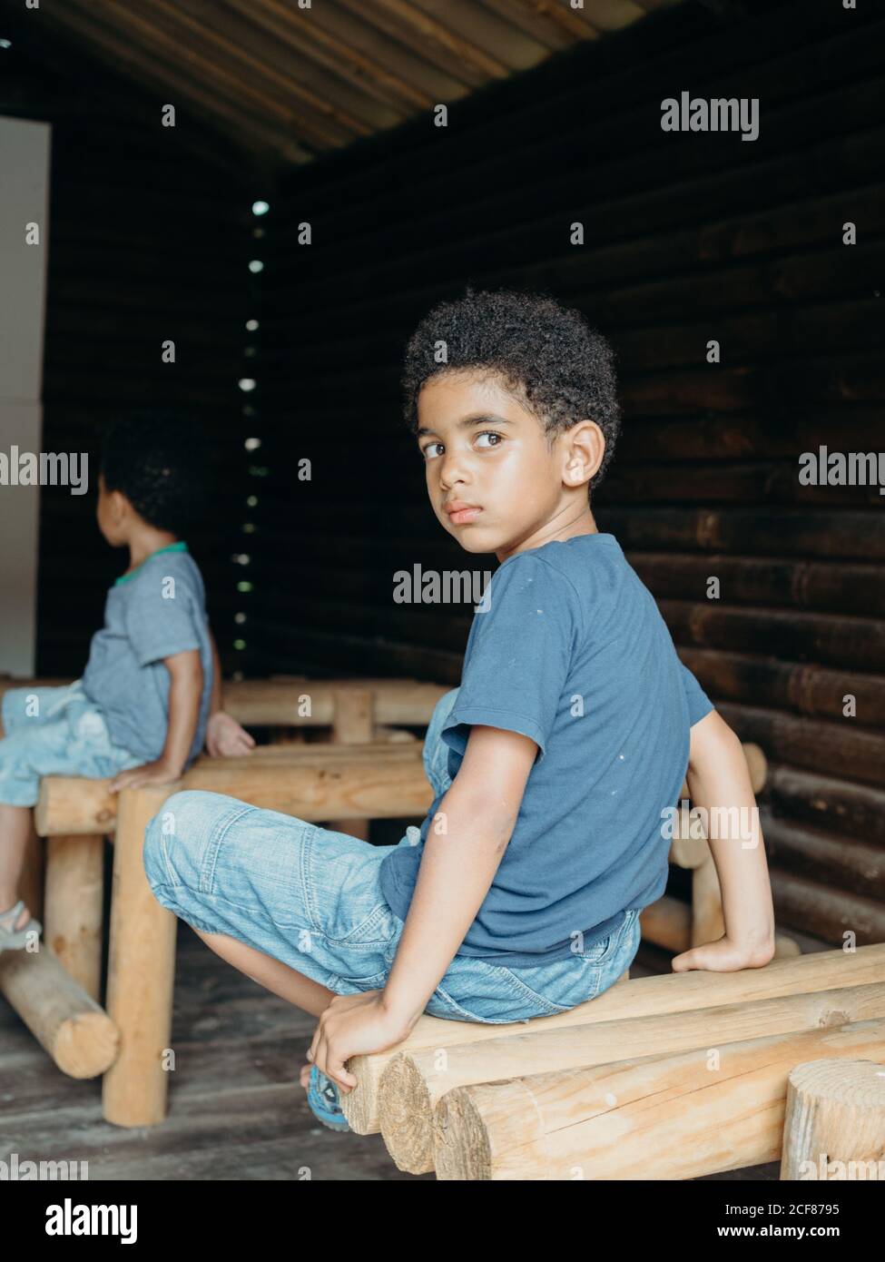 Cute African American boy in casual outfit looking over shoulder while sitting on wooden structure inside lumber building Stock Photo