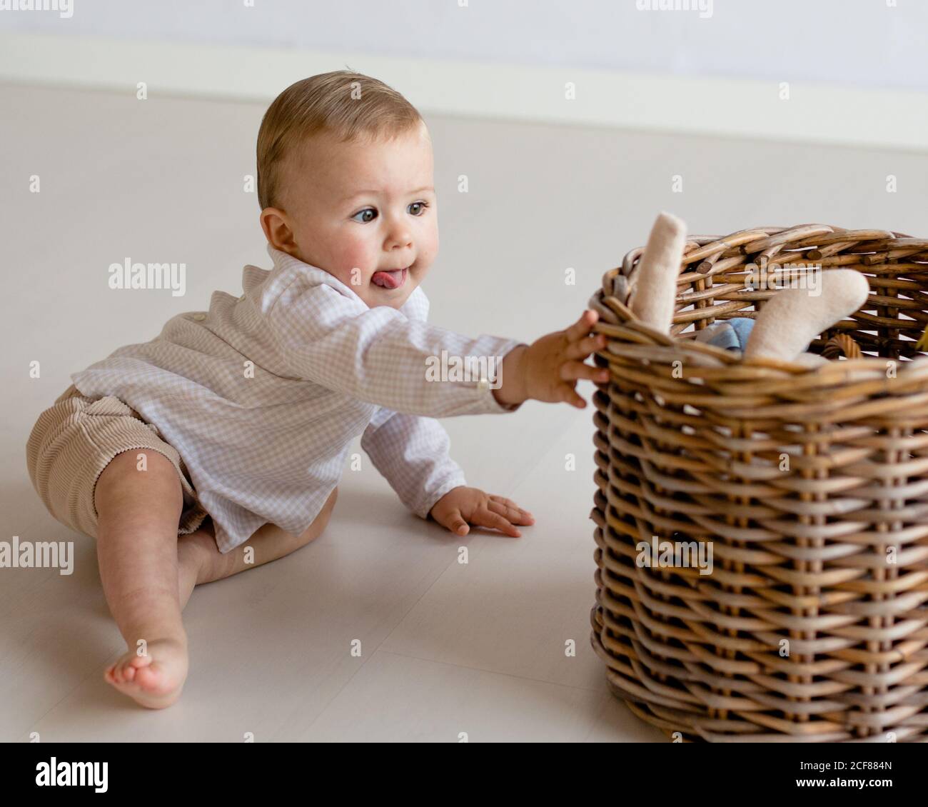 Curious little kid in casual wear sitting barefoot on floor at home and taking plush toys from wicker basket Stock Photo