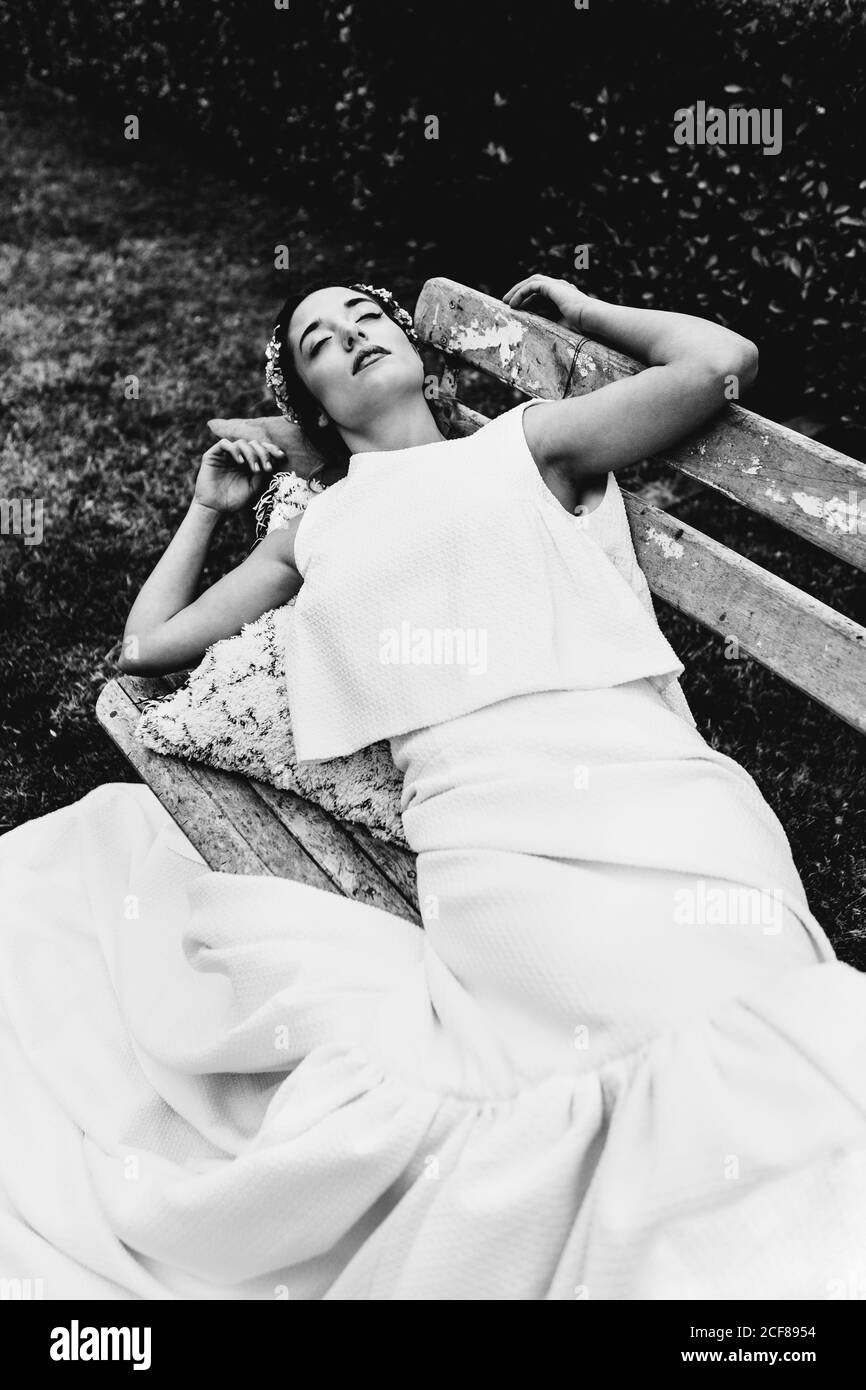 High angle of young female in white dress lying on bench and sleeping during wedding ceremony in park Stock Photo