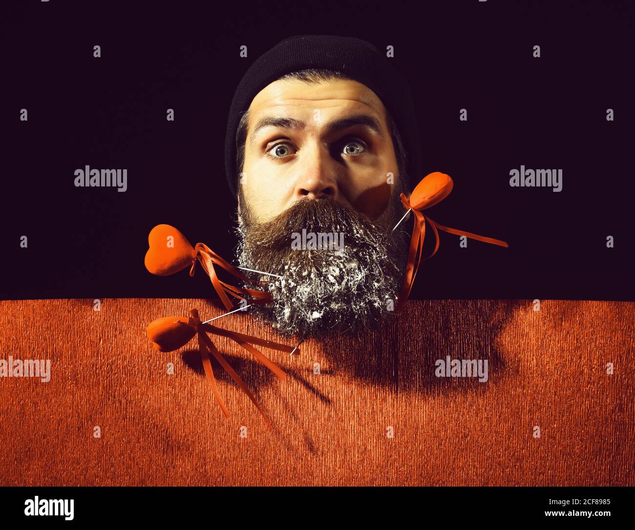 handsome bearded man or guy in winter hat with fashionable mustache on surprised face and snow in beard with decorative valentines hearts on sticks near red paper, on black background, copy space Stock Photo