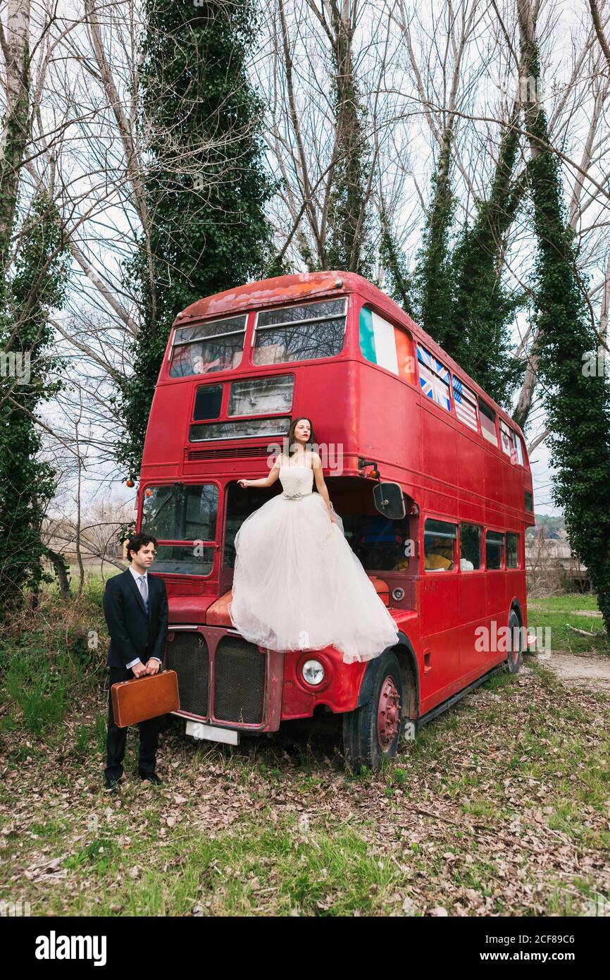 Bride in wedding dress sitting on hood of old double decker bus ...