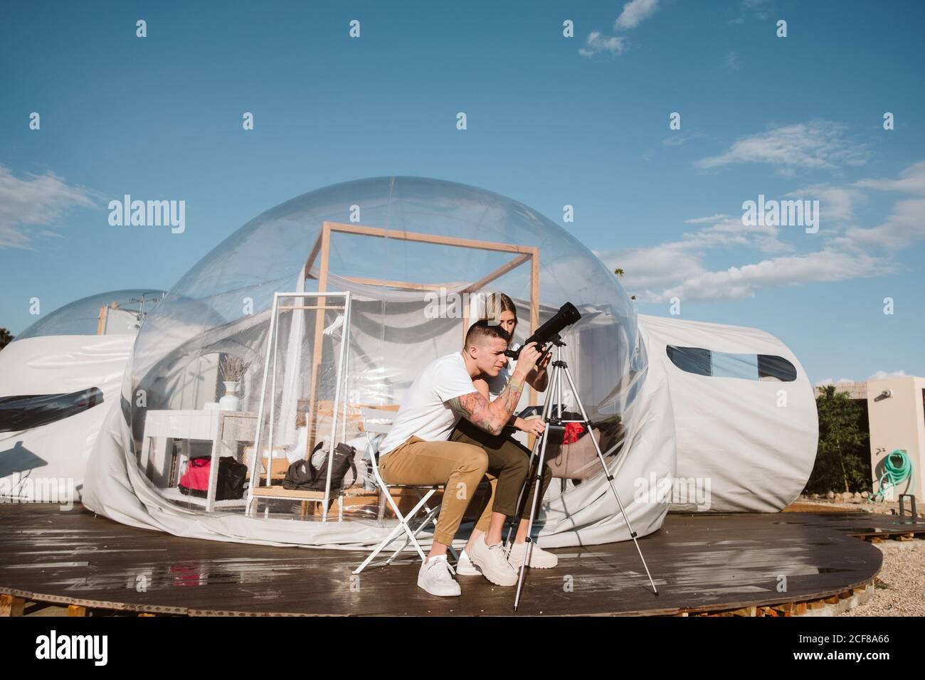 Stylish Woman sitting on boyfriend laps and looking through telescope at sky near bubble hotel Stock Photo