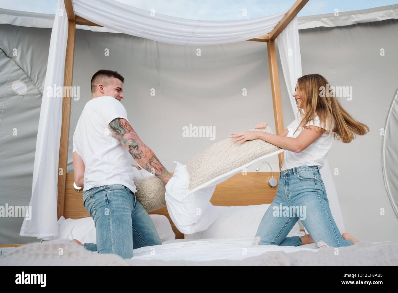 Cheerful young couple having fun during pillow fight on bed in big tent with transparent roof Stock Photo