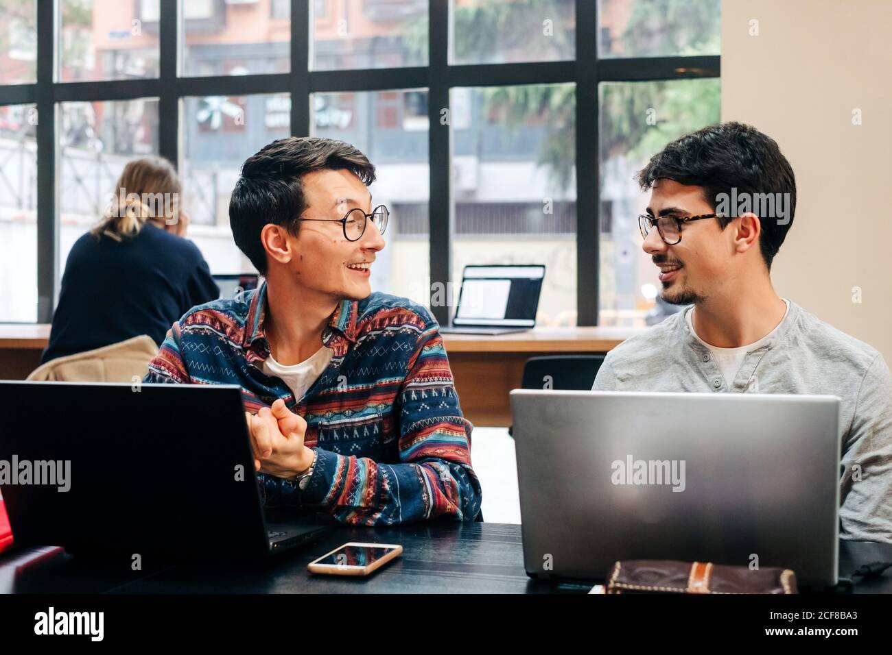 Positive coworkers in casual clothes and eyeglasses sitting at desk and working on project on laptops during workday in modern office Stock Photo