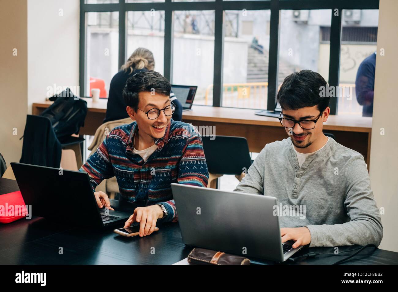 Positive coworkers in casual clothes and eyeglasses sitting at desk and working on project on laptops during workday in modern office Stock Photo