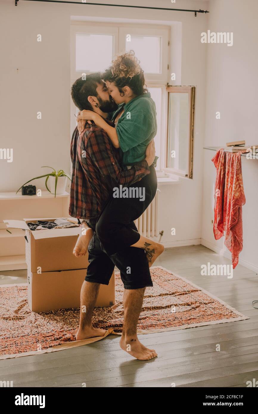 Side view of affectionate couple cuddling and front carrying while kissing beside stack of cardboard boxes at home Stock Photo