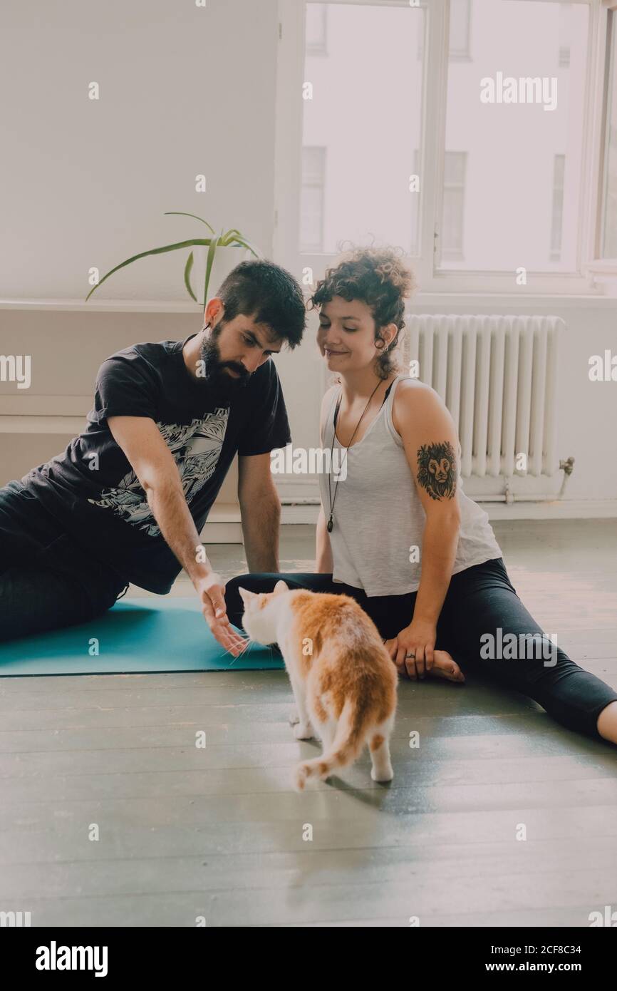 Side view of tender couple embracing while sitting on floor next to ginger cat at home Stock Photo