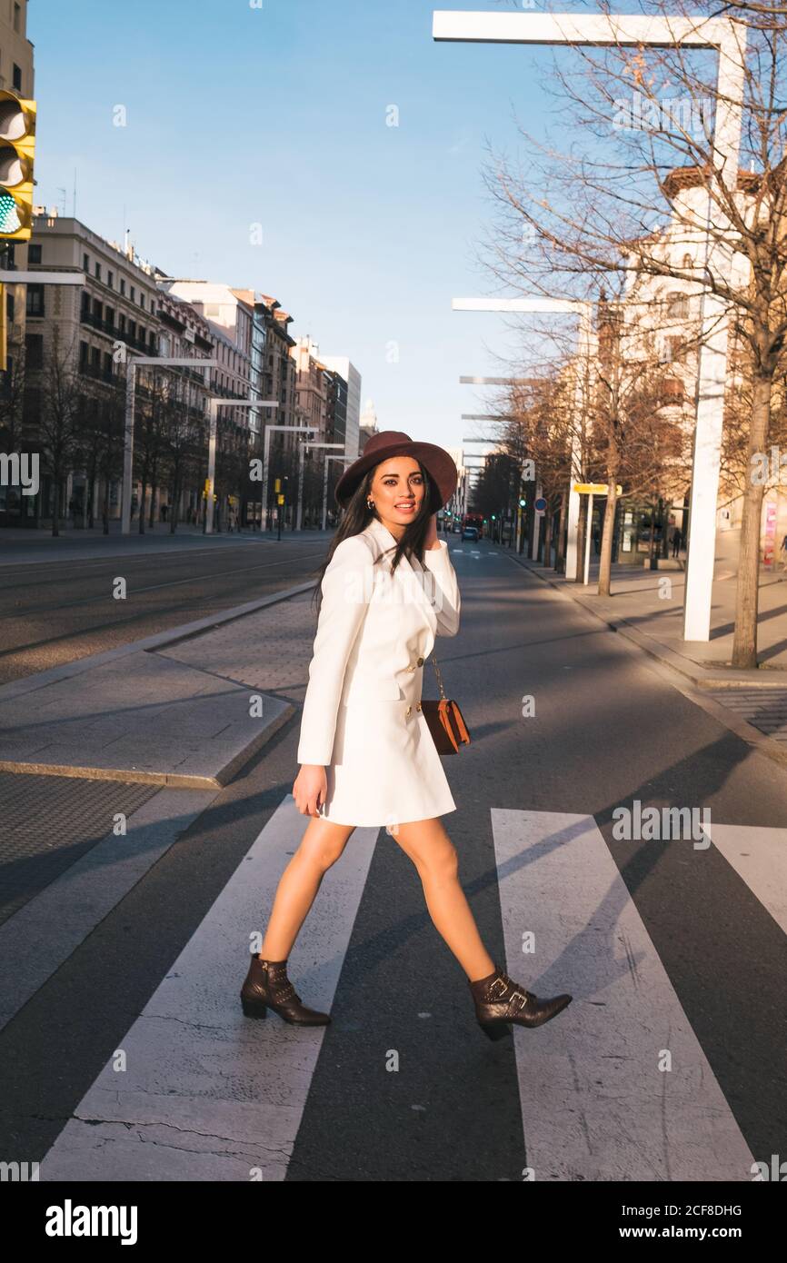 Full length side view of stylish young lady in white coat and trendy hat crossing street and looking at camera while walking in city in sunny spring day Stock Photo
