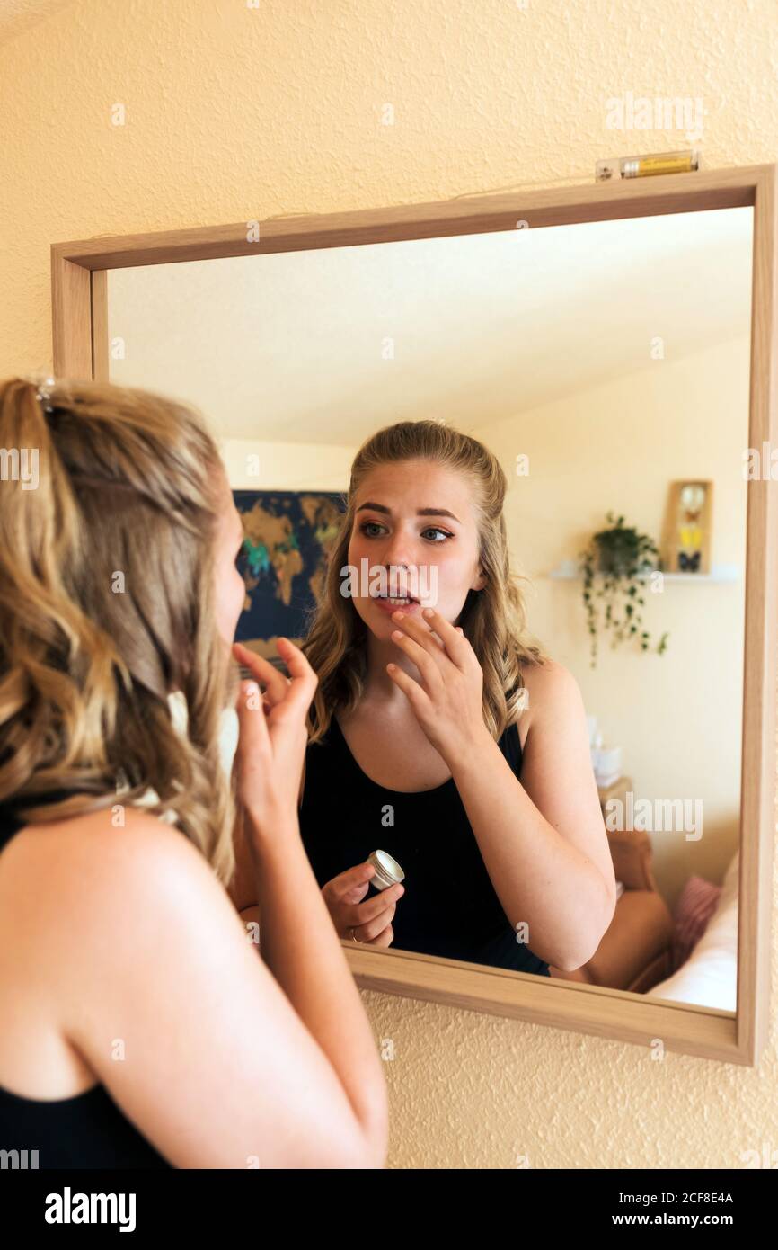 Young female in casual wear standing in front of mirror and applying lip balm while doing makeup at home Stock Photo