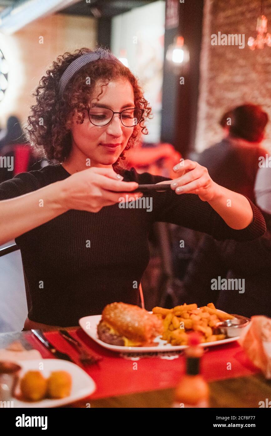 Young lady with curly hair using smartphone to take picture of burger and french fries while sitting at cafe table Stock Photo