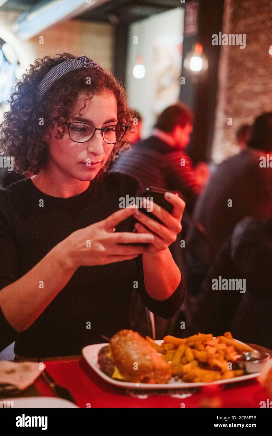 Young lady with curly hair using smartphone to take picture of burger and french fries while sitting at cafe table Stock Photo