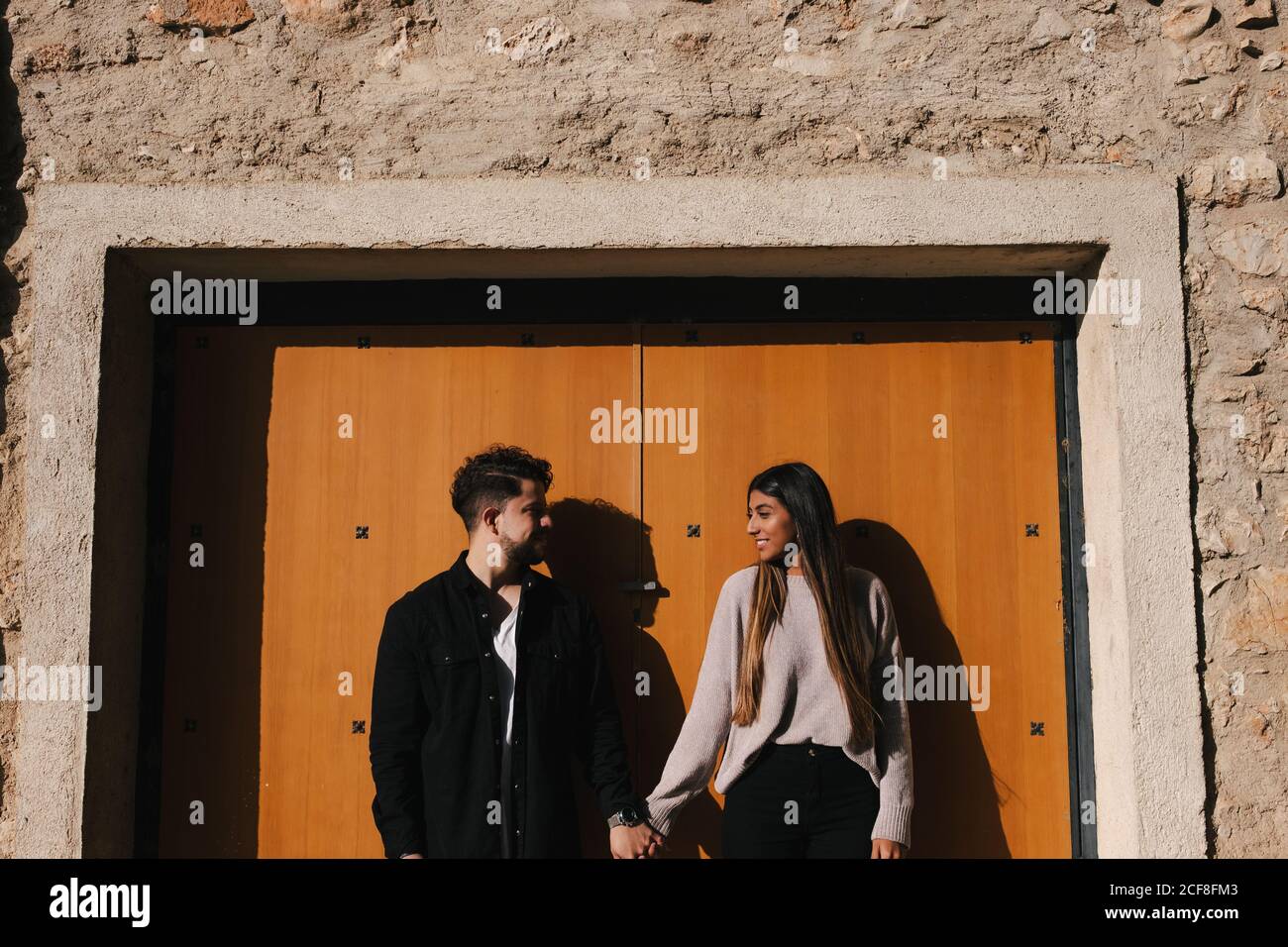 Smiling young ethnic couple in casual clothes holding hands and looking at each other with love while standing against entrance of aged stone building Stock Photo