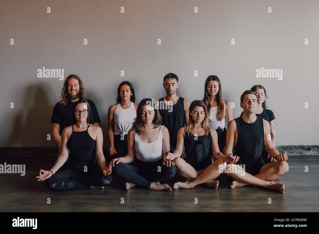 High angle of group of young serene women and men in sportswear with eyes closed and Gyan Mudra relaxing and meditating while sitting in padmasana position against white wall in contemporary yoga studio Stock Photo