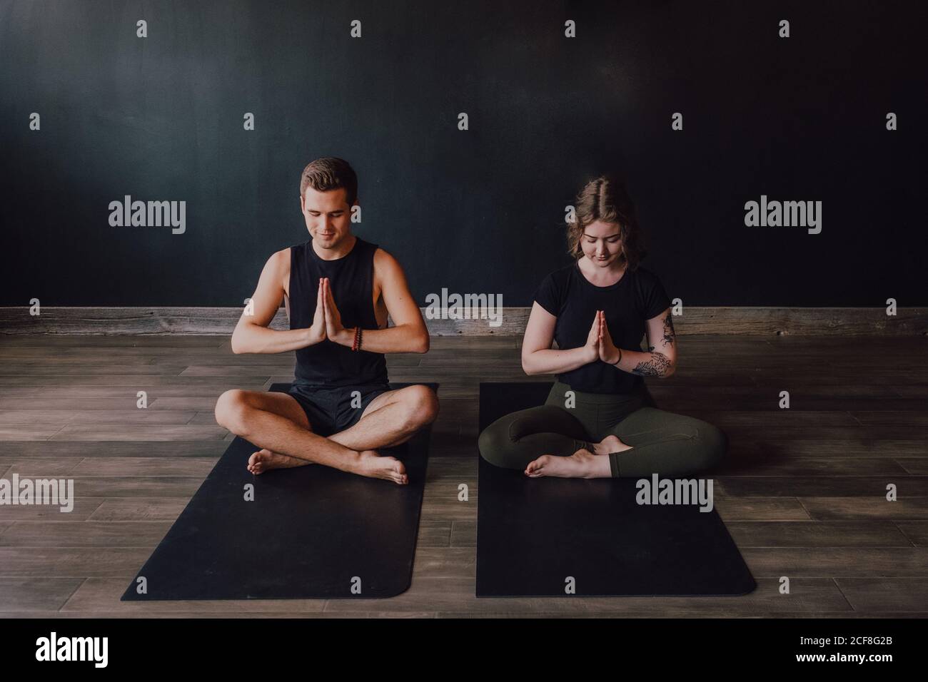 Calm young Woman and man in sportswear with eyes closed and legs crossed meditating together while sitting in padmasana position against back wall in contemporary yoga studio Stock Photo