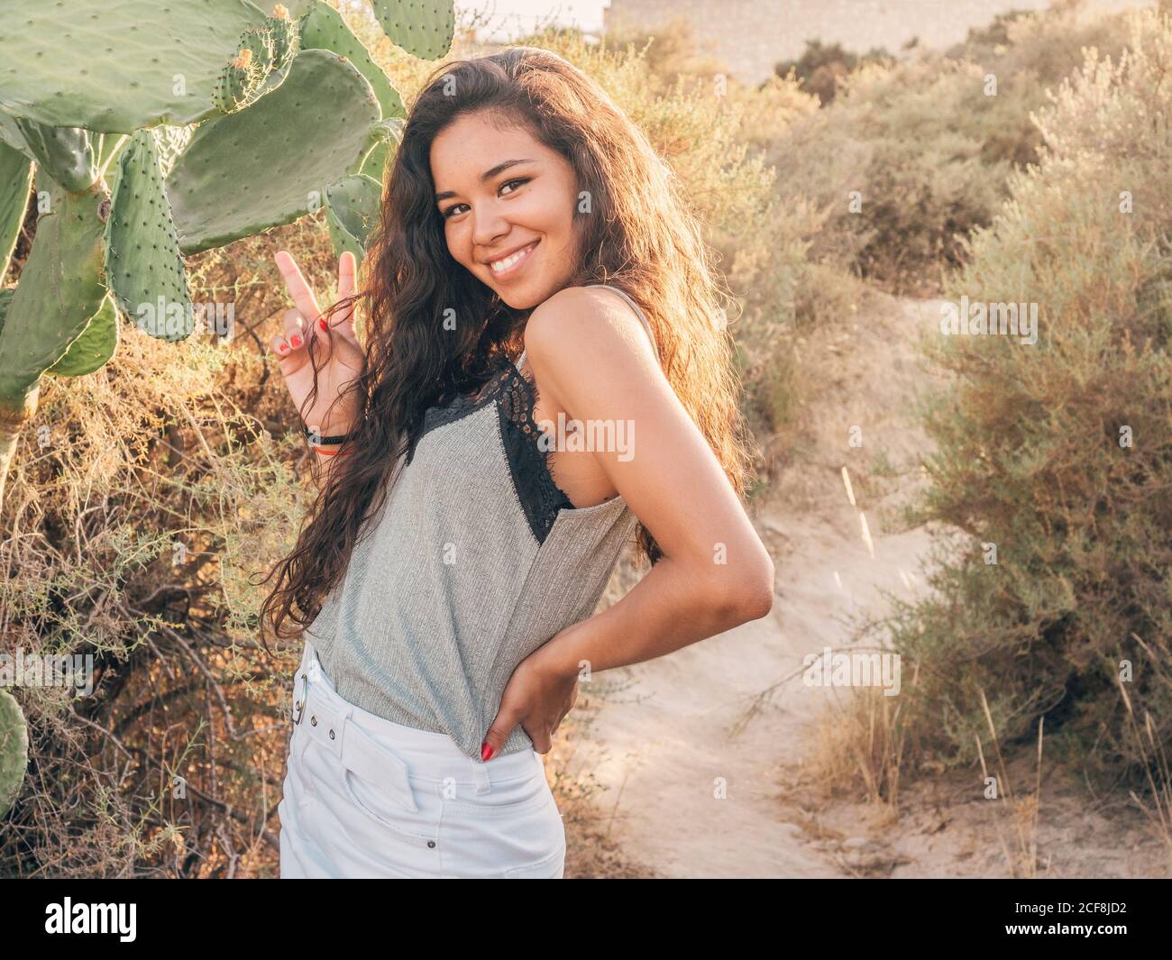 Smiling cheerful casual ethnic Woman standing beside cactus on sandy pathway amongst dry bushes in sunlight Stock Photo
