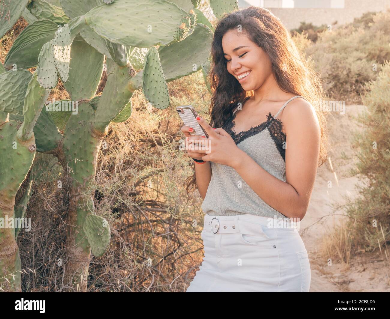 Smiling cheerful casual ethnic Woman browsing on smartphone while standing beside cactus on sandy pathway amongst dry bushes in sunlight Stock Photo