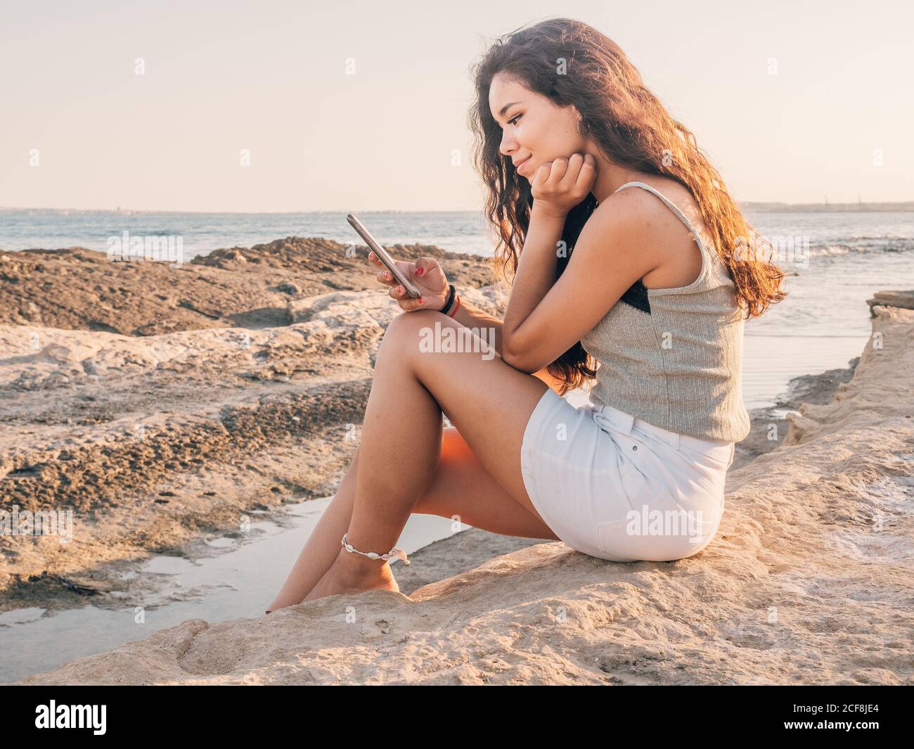 Side view of happy smiling casual ethnic Woman browsing on smartphone while sitting on seashore at sunset Stock Photo