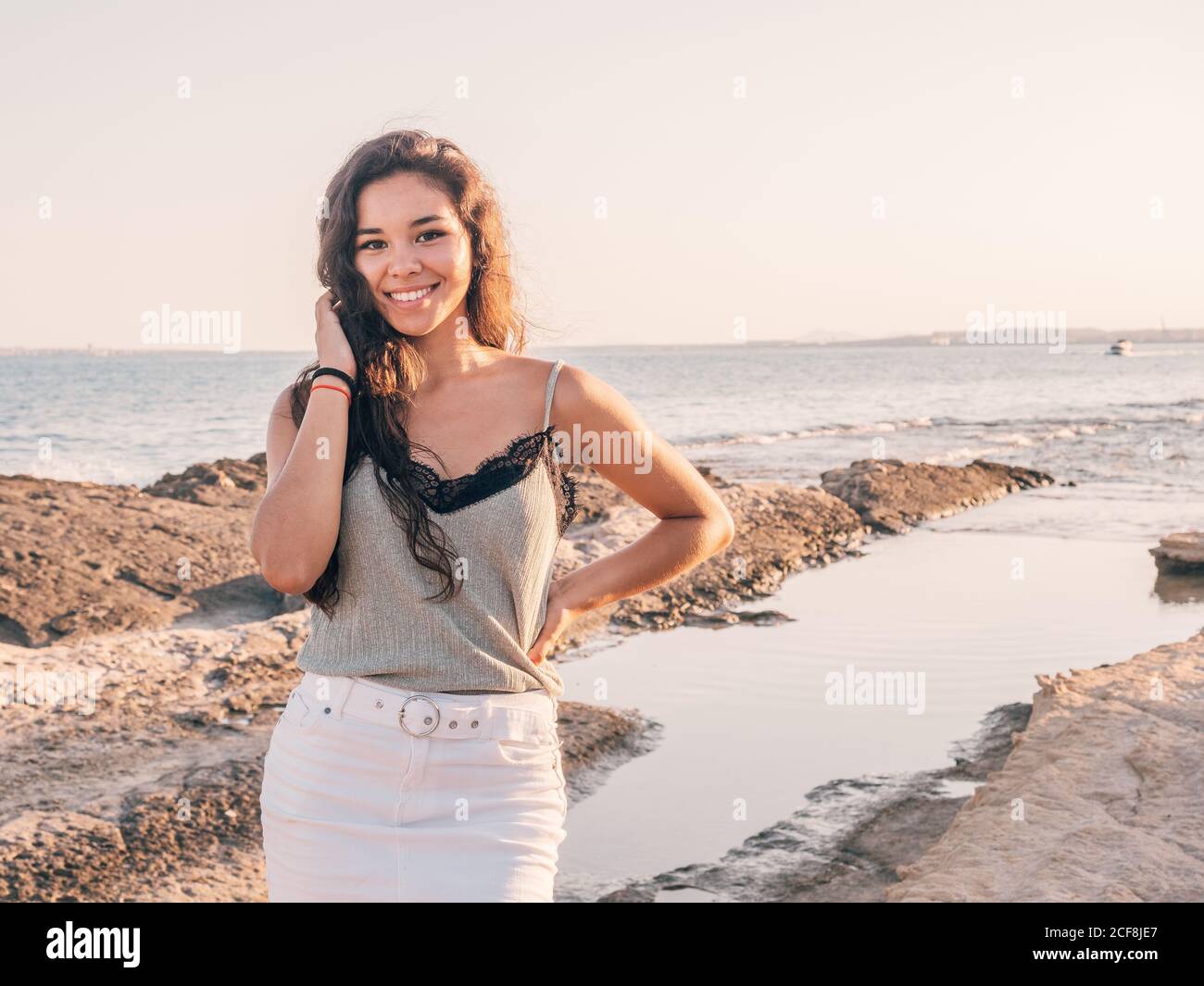 Smiling content casual ethnic Woman standing in beach beside coastline in sunlight Stock Photo
