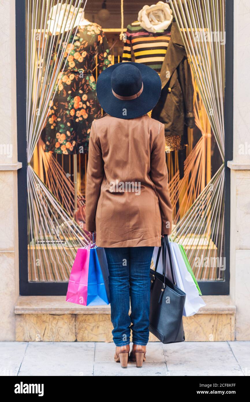 Back view of unrecognizable lady in stylish hat and brown jacket looking through shop window on fashionable dress with roses at city street Stock Photo