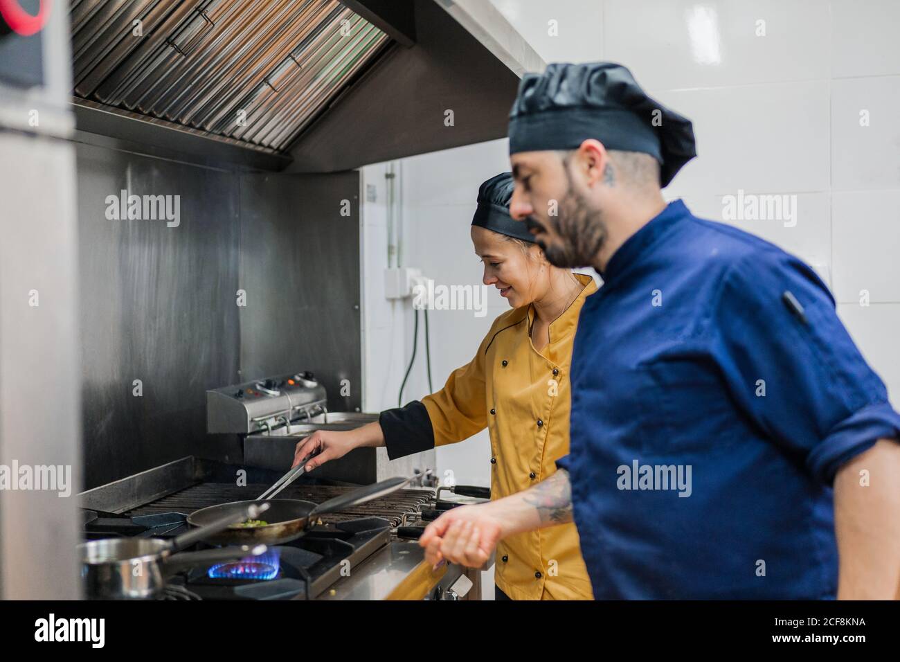 Side view of male chief chef watching young female assistant frying food while working together in professional kitchen Stock Photo