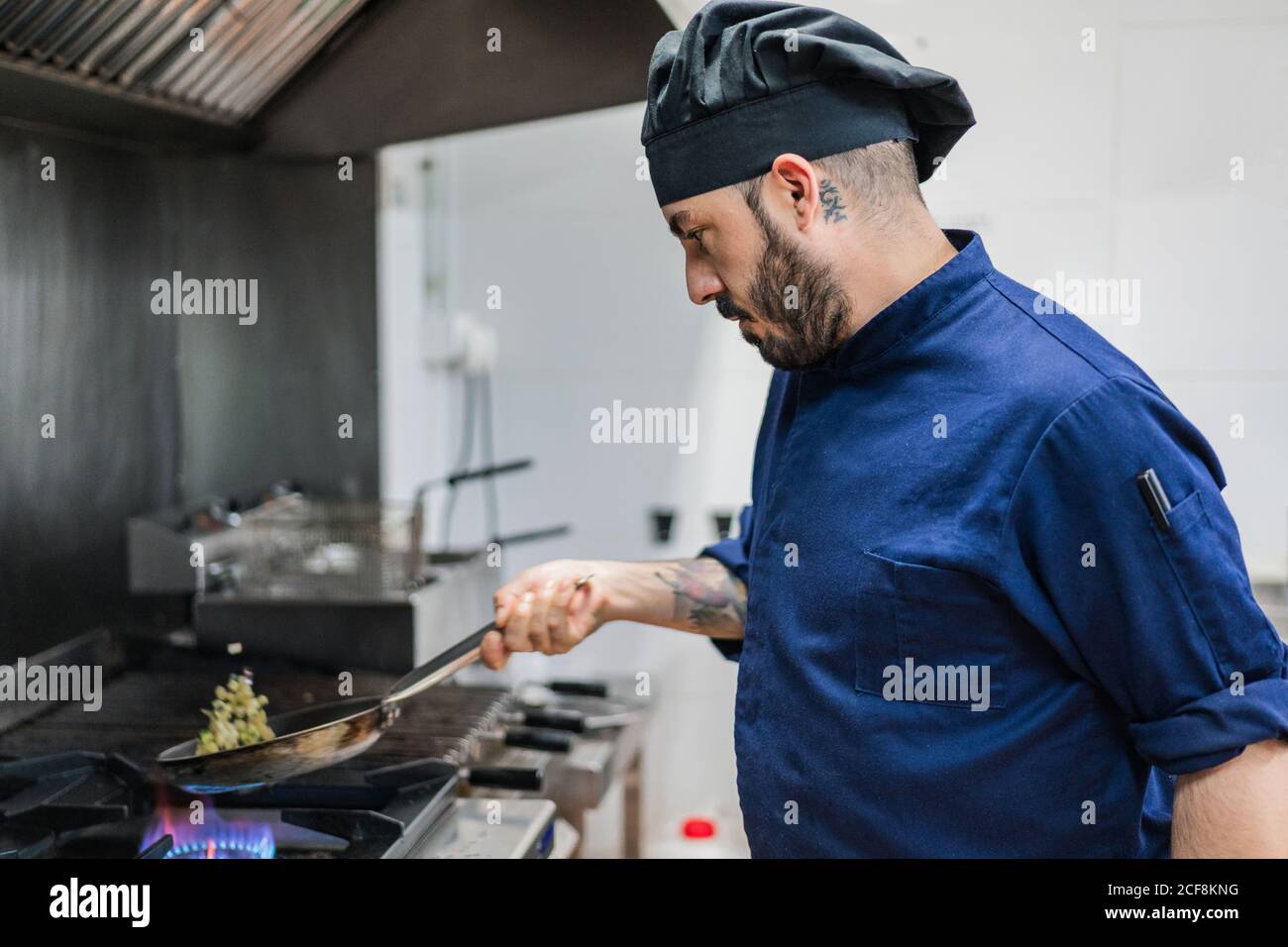 Side view of male cook in black hat shaking frying pan while cooking