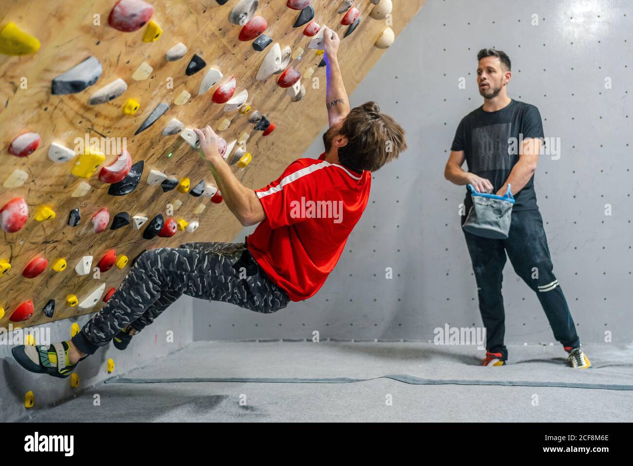 Side view of male athlete in sportswear climbing on grips of steep wall while male friend supporting with bag of talcum in gym Stock Photo