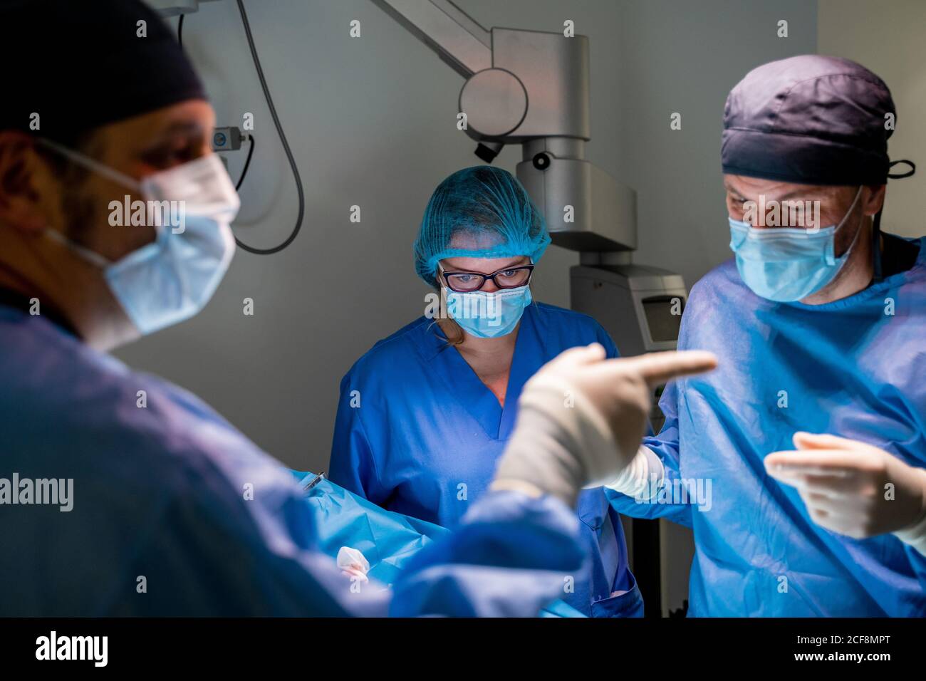 Pensive surgeon in uniform checking patient and making decision while coworker having consultation with colleagues during surgery in operating room of contemporary hospital Stock Photo
