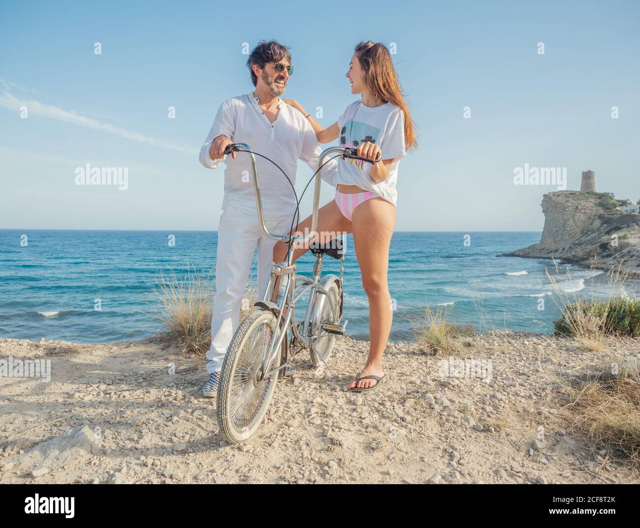 Stylish man and young Woman standing at seashore with bicycle enjoying amazing seascape in bright day Stock Photo