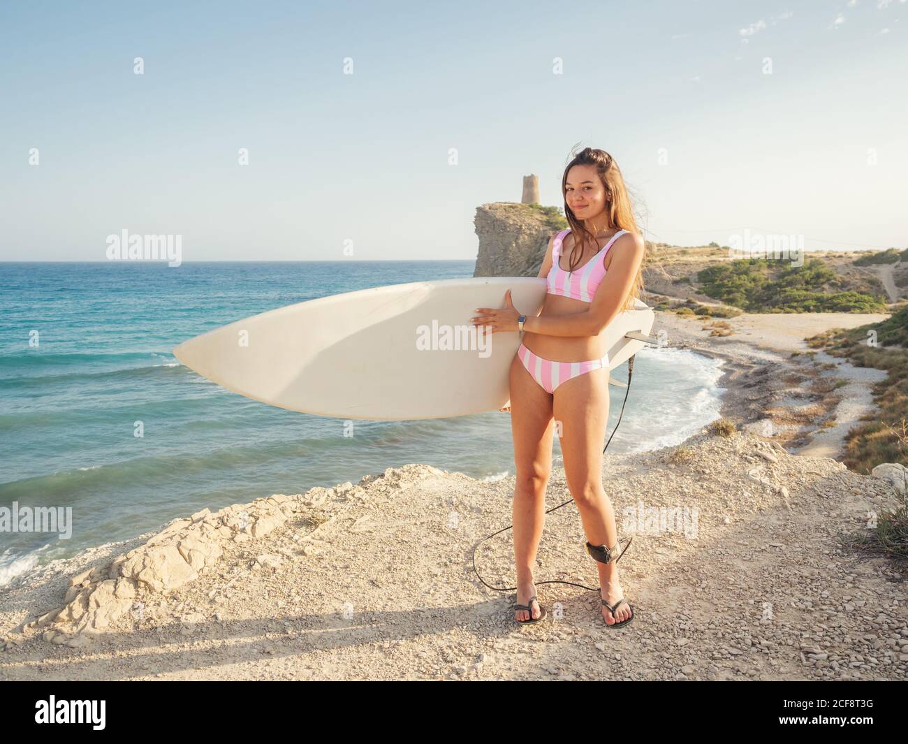Athletic tanned Woman going to surf raising surfboard high and looking at camera Stock Photo