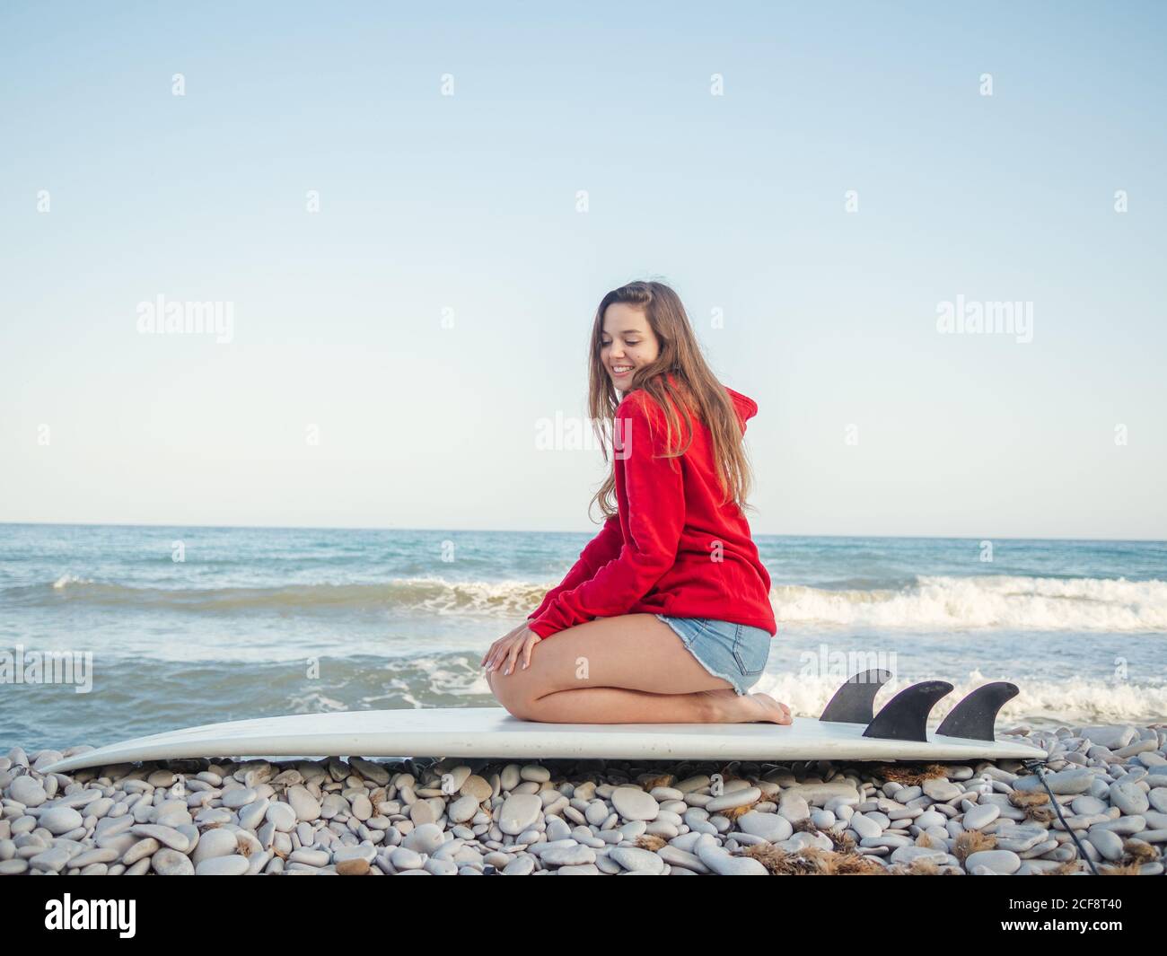 Side view of attractive smiling long haired Woman in red hoodie enjoying sitting on surfboard on stony seaside Stock Photo