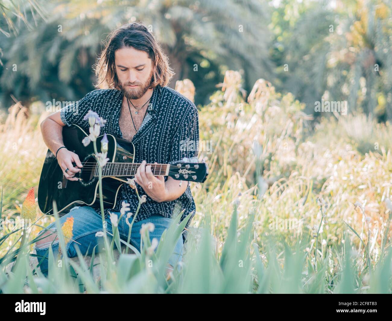 Pensive bearded hipster man sitting in meadow playing guitar Stock Photo