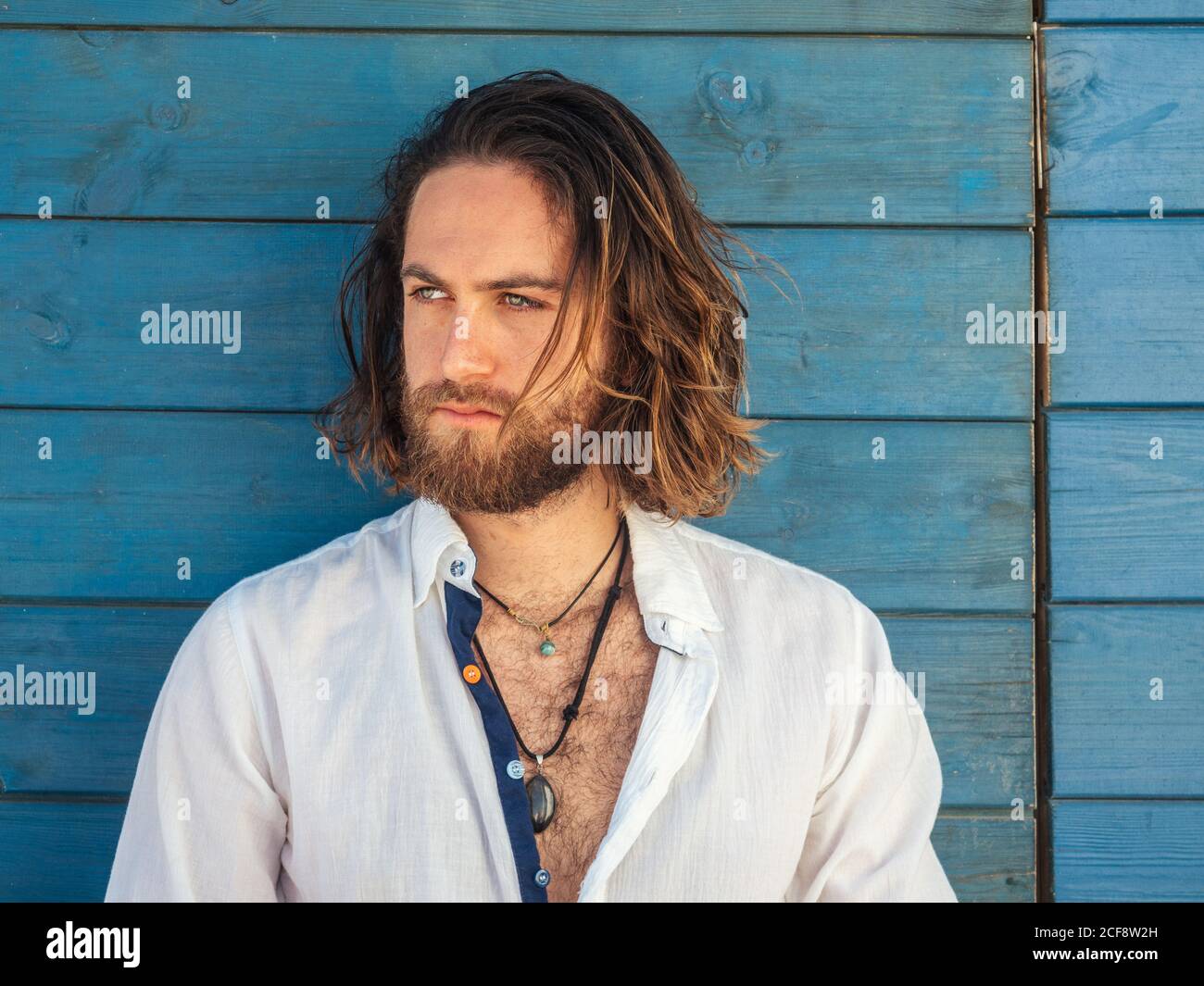 Confident bearded man in casual outfit and cheerfully smiling while sitting against blue wall on resort Stock Photo