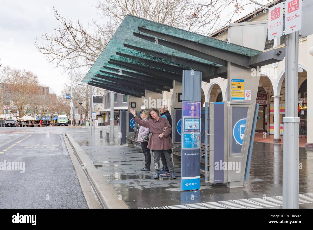 On a rainy day a woman waves down an oncoming bus at a bus stop in central Canberra City, ACT, Australia Stock Photo