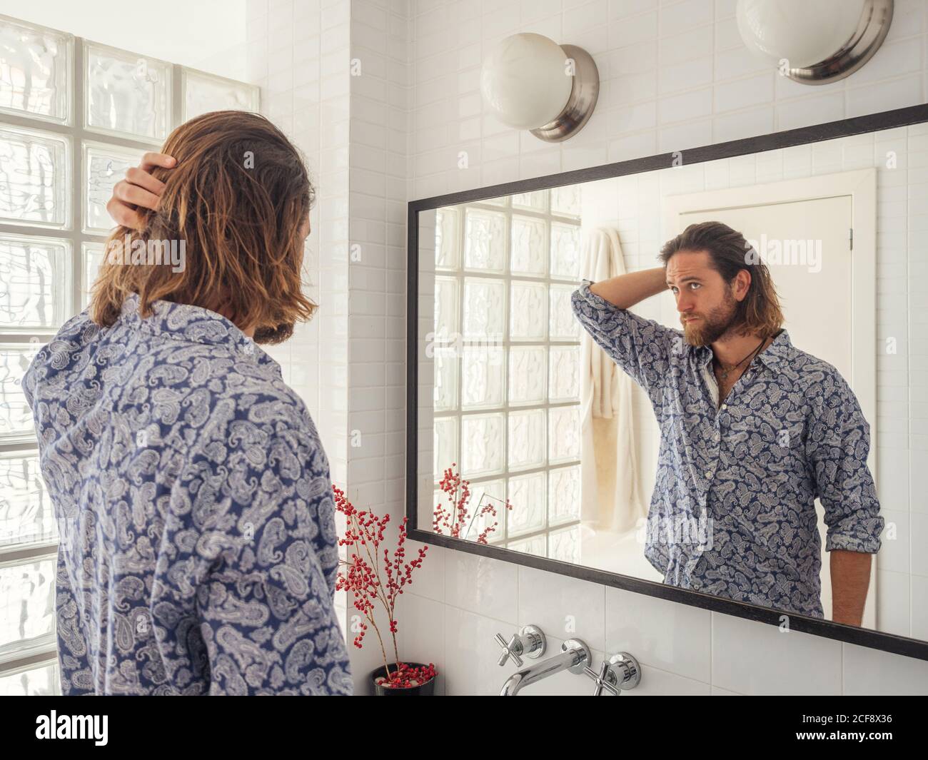 Back view of cool adult man fixing hair in standing in light bathroom in front of mirror Stock Photo