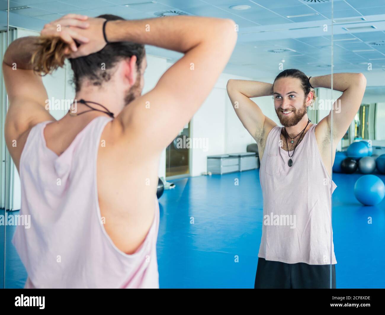 Cheerful young bearded man collecting hair in tail while standing in front of mirror in spacious gym Stock Photo