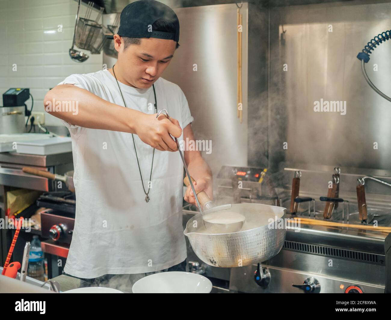 Young man in black cap pouring hot broth from saucepan for Japanese dish called ramen in Asian cafe Stock Photo